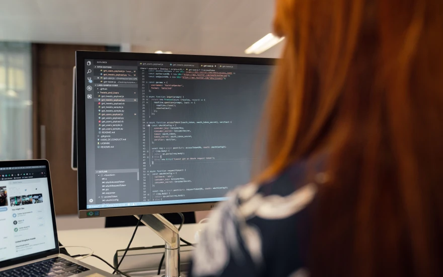 Someone at a large monitor filled with a code editor and an open file tree, a laptop beside it on the desk.