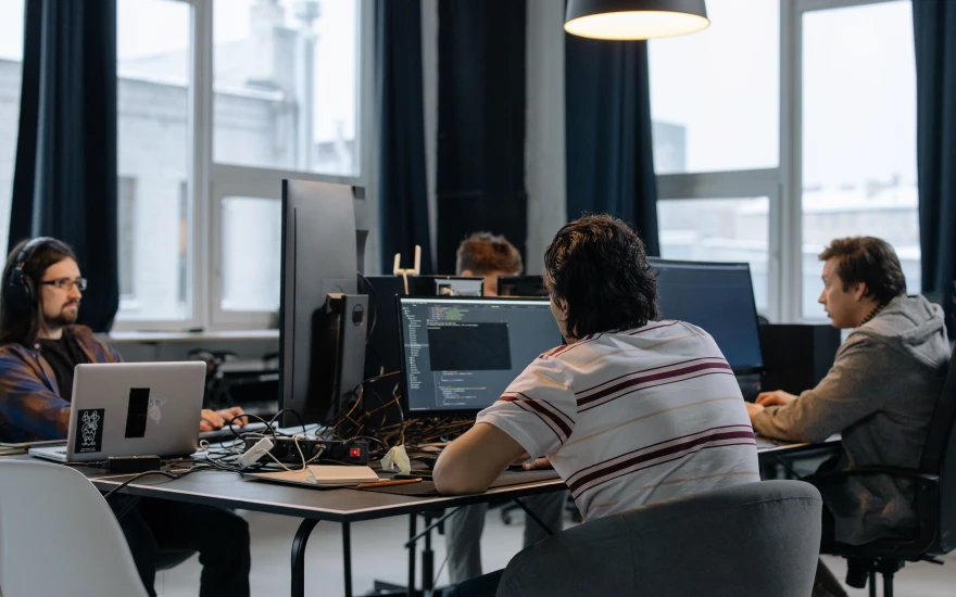 Four people working at adjoining desks in a bright office, one on a laptop and one at a large monitor showing a code editor.