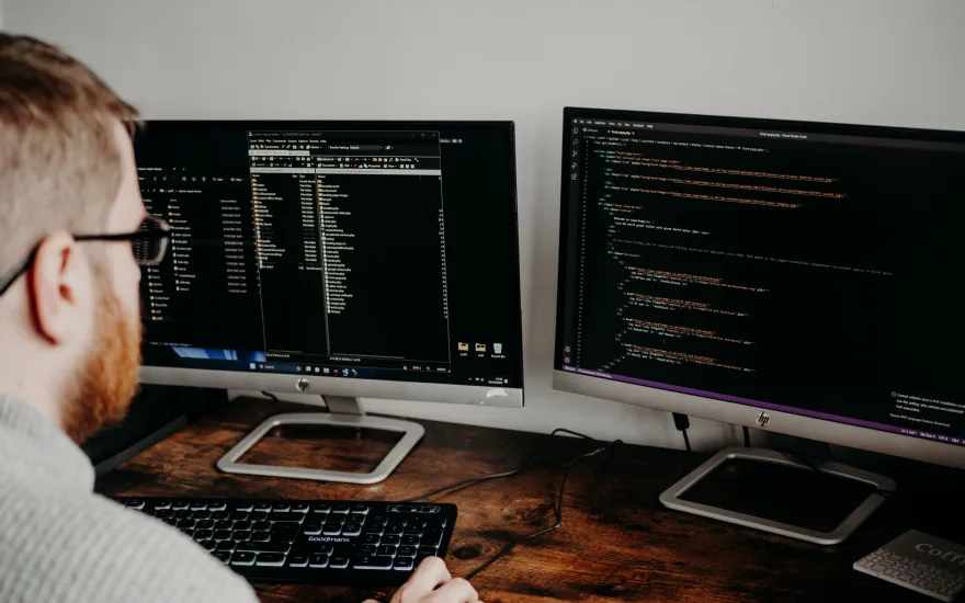 A developer at a wooden desk working across two monitors, one showing a file manager and the other a code editor.